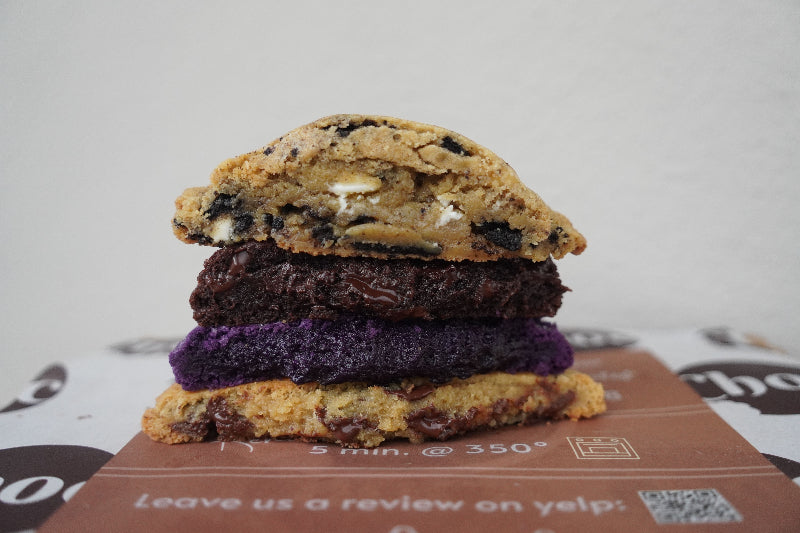 CHOC Cookies' signature four-cookie stack featuring vibrant purple Ube, rich Cookies & Creme with Oreo pieces, dark chocolate with melted center, and classic chocolate chip cookies split to showcase each unique texture and filling, professional bakery photography
