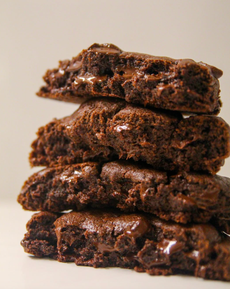 ecadent triple chocolate fudge cookies stacked and split to reveal gooey melted chocolate center, showcasing rich brownie-like texture with glossy chocolate chunks throughout, professional bakery photography - better than crumbl